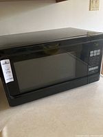 Front view of the black microwave showing the door with a large viewing window and lot tag on the left corner.
