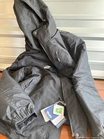 Full view of black winter coat with visible zippered pocket and attached tags on sleeve area.