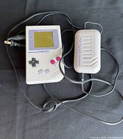 Front view of the original Nintendo Game Boy handheld gaming console with charger attached, showing control buttons and screen.