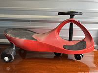 Side profile of red Playsma ride-on car showing seat, body shape, rear wheels, and front steering wheel column.