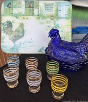 Photo showing six colored shot glasses with gold accents, blue glass rooster dish, and farm-themed glass cutting board