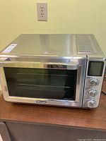 Side-angle view of the stainless steel toaster oven placed on a wooden surface with clear glass door and visible brand logo on bottom center.