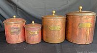 Four vintage copper canisters with visible patina and labeling plates in a row against black backdrop.