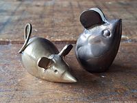 Two brass mouse miniature figurines shown from an angle on a wooden surface. One figurine has a smooth polished finish with small ears and coiled tail. The other has a matte finish with large rounded ears and rounded body.
