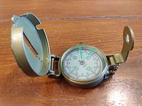 Engineer compass open on wooden surface, showing dial and brass casing with hinged lid.