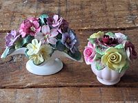 Photo shows two porcelain floral arrangements on a wooden surface: one with varied flower colors on a cream crescent base, the other a pink round vase with roses.