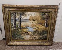 Framed landscape painting showing trees, stream, flowers in gold-tone painted frame on carpeted floor.
