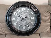 Photo of large round wall clock with black plastic frame and gear design on couch.