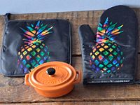 Photo of mini orange ceramic cocotte with lid, black oven mitt, and pot holder featuring colorful pineapple designs.