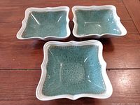 Photo showing three rectangular stoneware bowls with scalloped edges in teal green glazed interiors and white exteriors, arranged on wooden surface.