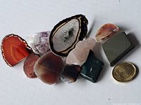 Multiple natural stone samples arranged on white background with a coin for scale, showing polished and rough stones including agate, amethyst, quartz, jasper, hematite