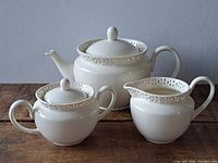Photo of cream colored ceramic teapot, sugar bowl, and creamer with lace-like decorative border on a wooden surface against gray background.