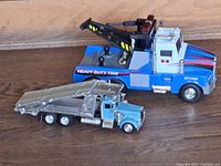 Two toy trucks on wood surface, one large blue tow truck with crane, smaller metallic loader truck in front