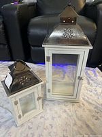 Two white lantern boxes side by side on fabric, showing metal peaked tops with floral design, glass sides and doors with metal latches.