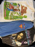 Three T-shirts laid out showing front graphics: white with alien safety education print, blue with Superman logo, black with portrait and text.