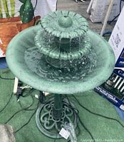 Electric plastic bird fountain operational and filled with water, showing water flowing from the top tier to lower basin on green indoor/outdoor carpet background.