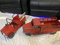 Two red metal trucks, one pickup style and one jeep style, shown side by side on a cloth with black leather couches in background.