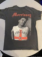 Front view of black Morrissey t-shirt with graphic of Morrissey wearing an England cross shirt and 'Morrissey' text above in red.