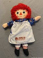 Raggedy Ann doll replica with red yarn hair, blue floral dress, and white apron with ARCS Mortgage logo.