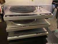 Stacked three vintage direct drive turntables with clear dust covers, placed on a table indoors.