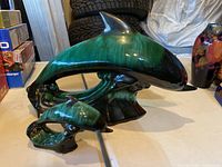 Photo showing two ceramic dolphin figurines made by Blue Mountain Pottery, green and black glazed with wave bases, placed on table with other items in background.