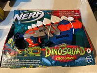 Front view of boxed Nerf DinoSquad Stego-Smash dart blaster showing the product and darts included.