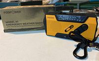 Front and side view of yellow and black Fospower Model A1 emergency weather radio sitting next to its original box.