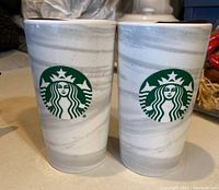 Front view of two Starbucks ceramic travel mugs showing green logo on white marble pattern.