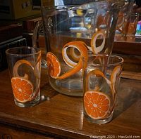 Clear glass pitcher and two glasses with orange slice and peel designs, shown on wooden surface with reflection in background mirror.