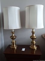 Two brass table lamps side by side on a wooden surface, showing overall size and condition including white fabric shades.