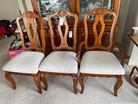 Three wooden dining chairs with upholstered light beige seats and carved backs, one with arms, set in front of a china cabinet.