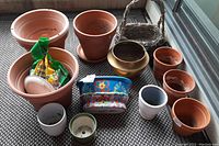 Photo showing 12 assorted garden pots in clay, ceramic, plastic, one brass-colored metal pot, 1 wicker basket, and opened bag of potting soil on patterned surface near glass door.