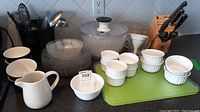 Shows black ceramic utensil holder with various metal and plastic utensils, clear glass salad plates stacked, white ceramic pitcher, white ceramic ramekins and bowls stacked, salad spinner, and green cutting board.