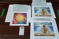 Three art prints with Certificates of Authenticity, one by Alfred Gockel showing a stylized yellow tulip, two by Peter Nixon showing a woman sitting by water, all on a wooden surface.