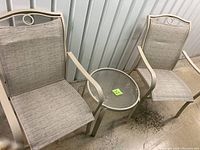 Two beige outdoor chairs and a round side table arranged with visible metal frames and woven fabric seats and backs