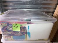 Transparent plastic storage bin filled with games, puzzles, and coloring items with a lot number sticky note