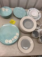 Large lot of blue and white casual dinnerware displayed on table including plates with shell motifs, bowls, mugs, and luncheon plates.