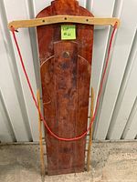 Photo of the full sled standing upright against a wall showing the wooden body, curved handle with red rope, and metal runners.