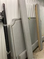 Side view of the upholstered king headboard leaning upright showing light gray fabric upholstery and metal bedframe components leaning beside it