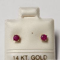 Front view of both ruby stud earrings mounted on white textured display card with '14KT GOLD' in gold lettering.