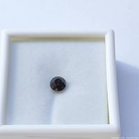 Close-up photo of a 0.6 carat round cut black diamond loose gemstone placed on white cushioned padding inside a square white jewelry display box.