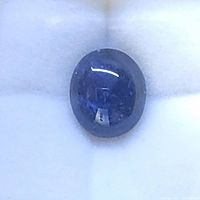 Close-up of oval cabochon sapphire gemstone resting on a white cushion background, showing blue hue and internal inclusions.