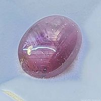 Close-up of oval cabochon star ruby gemstone showing pinkish hue, surface reflection spot, and internal star effect with visible inclusions.