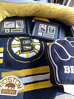 Framed memorabilia with two Boston Bruins player cards and nameplates, a Boston Bruins rug with logo, and a black/yellow pillow with Bruins text, all placed on a couch.