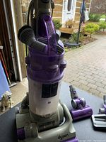 Full view of purple and gray Dyson upright vacuum cleaner with a transparent dust bin and front suction opening, standing on a wooden floor.
