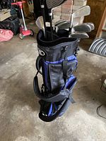 Photo of a black and blue golf bag standing upright in a garage area, showing side pockets and strap handles