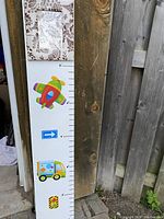 Full view of the height measurement chart placed against a wooden fence, showing colorful airplane, truck, and traffic light graphics beside ruler markings.