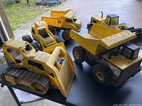 Five yellow kids heavy equipment toys on a black table at an angle showing different sides. Vehicles include large bulldozer with tracks, dump trucks, and loaders.