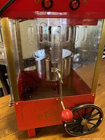Front view showing red metal body, glass panels, metal kettle with stirrer inside, and wheels.