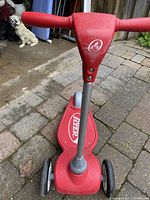 Red Radio Flyer kids scooter with three wheels shown from front and top angle outdoors on brick and stone floor.